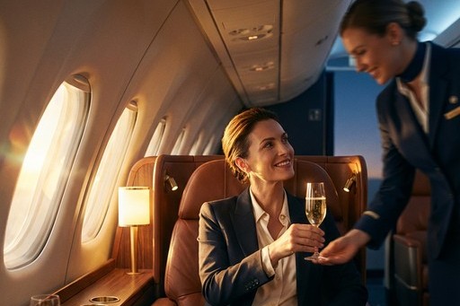 A seated passenger in a premium cabin is handed a glass by crew, low evening light through the window.